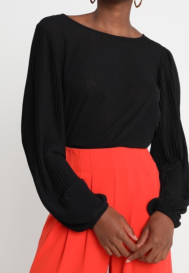 Mustapaita, jossa on pitkät pleatatut hihat ja leveä kaula-aukko, yhdistettynä kirkkaanpunaisiin korkeavyötäröisiin housuihin, joissa on terävät pleatit.