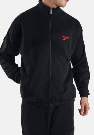 Mann trägt eine schwarze Reebok-Jacke mit durchgehendem Reißverschluss, strukturierten diagonalen Paneelen und rotem Logo auf der Brust, Hände entspannt an den Seiten.