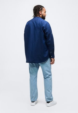 Homme aux cheveux tressés portant une chemise à manches longues bleu foncé, un jean bleu clair et des baskets blanches, debout de dos sur un fond uni.