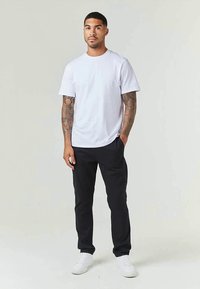 Homem de pé a usar uma t-shirt branca, calças pretas, ténis brancos, com tatuagens em ambos os braços, contra um fundo claro e simples.