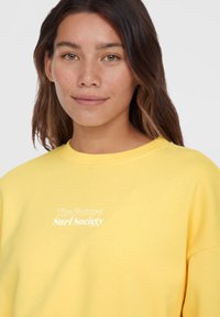 Žltá mikina s okrúhlym výstrihom, s bielym textom "The Future Surf Society." Mäkká tkanina, voľný strih, mierne oversize dizajn.