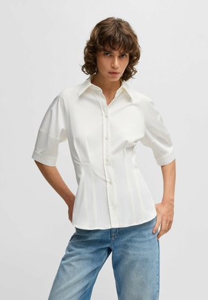 Camicia bianca a maniche corte, vestibilità aderente, con pieghe sottili e una texture del tessuto liscia. Abbinata a jeans azzurri.