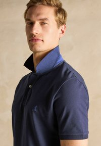 Tumesinine polo särk tekstuurse disainiga, millel on kontrastne sinine triibuline krae ja väike brodeeritud logo rinnal.