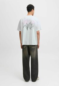 Světle zelené oversized tričko s růžovým a černým květinovým vzorem na zádech, spojené s tmavými kalhotami s širokými nohavicemi a neutrálními teniskami.