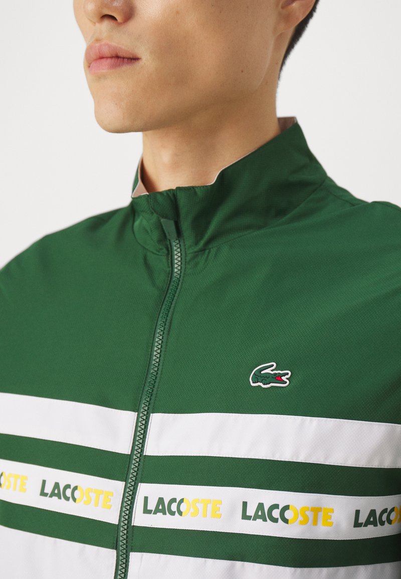Veste zippée verte avec des rayures blanches et une inscription 'LACOSTE' jaune. Dotée d'un col montant et d'un petit logo crocodile vert sur la poitrine.