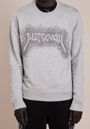 Personne portant un sweat-shirt gris clair avec un motif clouté noir formant un motif abstrait sur la poitrine.