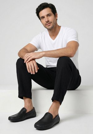 Mand med mørkt hår sidder på en hvid blok iført en hvid t-shirt, sorte bukser og sorte slip-on sko mod en ensartet lys baggrund.