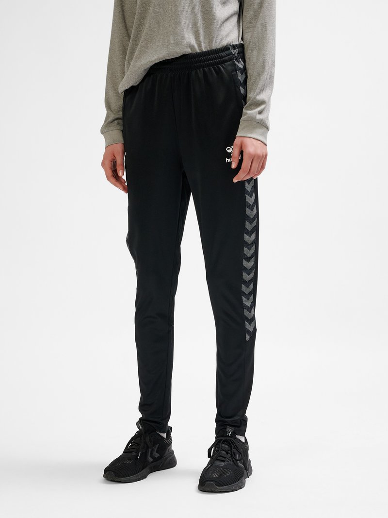 Person iført sorte taperede træningsbukser med sideløbende chevronstriber, sorte sneakers og en grå langærmet trøje stående mod en hvid baggrund.