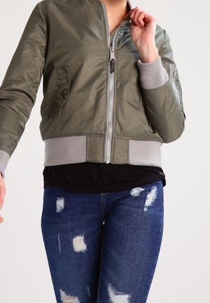 Personne portant un blouson aviateur vert olive avec des poignets et une ceinture gris, un t-shirt noir et un jean bleu déchiré sur un fond uni.