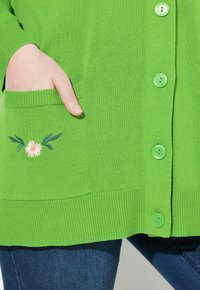 Grön cardigan i strukturerad stickad tyg, med knappfram, ficka med rosa blomstert broderi och ribbade ärm och fåll.