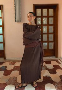 Pitkään ruskeaan plissee mekkoon leveät hihat ja yhteensopiva vyö, jossa on teksturoitu kangas, joka laskeutuu lattiaan ja sandaalit.