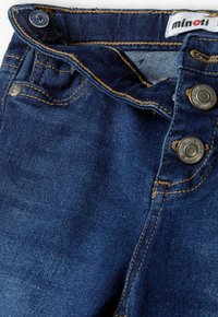 Modre denimske hlače z zlatimi šivi, dvema kovinskima gumboma na pasu in oznako "minoti" blizu vrha. Tekstura tkanine.
