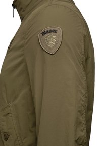 Giacca verde oliva con una patch testurizzata sulla spalla con la scritta "Blauer", chiusura con zip e dettagli delle tasche laterali.