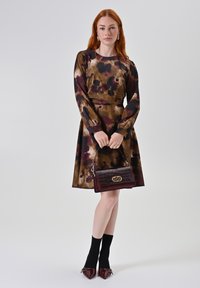 Jurk met lange mouwen in een bruine en maroon bloemenprint, gecombineerd met een bordeaux-rode gestructureerde handtas en zwarte enkelsokken. Spitse schoen.