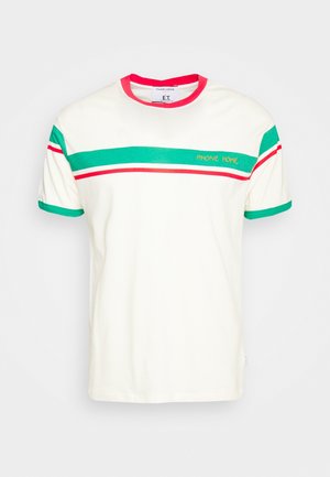 Osoba ubrana w biały T-shirt w zielone i czerwone paski z napisem „PHONE HOME”, brązowe spodnie i białe masywne trampki.