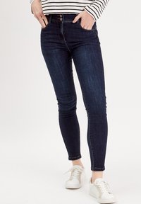 Jeans skinny bleu marine avec taille haute, cinq poches et texture lisse. Associés à des baskets blanches avec des accents beiges.