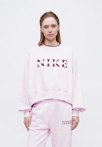 Svetloružová mikina Nike s výrazným burgundským logom, s voľným strihom a širokými rukávmi, v kombinácii s nohavicami na cvičenie v rovnakom odtieni.
