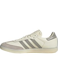 Zapatilla color crema con acentos de gamuza y cuero gris, tres rayas grises y suela de goma texturizada. Diseño de perfil bajo con cordones.