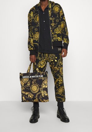 Mężczyzna noszący czarną i złotą kurtkę i spodnie w wzory, trzymający pasującą torbę z logo Versace Jeans Couture. Widoczne czarne buty.