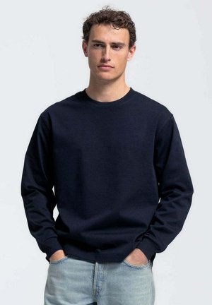 Jonge man met krullend haar, draagt een marineblauwe trui met ronde hals en lichtblauwe spijkerbroek, handen in de zakken, neutrale gezichtsuitdrukking.