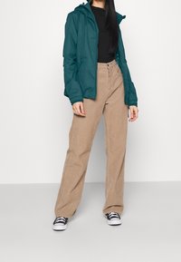 Žena v teplém modrém zipovaném svetru, černém tričku, béžových kordových volných kalhotách a černo-bílých teniskách stojící na bílém pozadí.