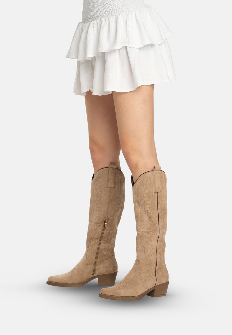 Mujer con botas vaqueras de ante marrón claro hasta la rodilla, con cremalleras laterales y falda corta blanca con volantes.