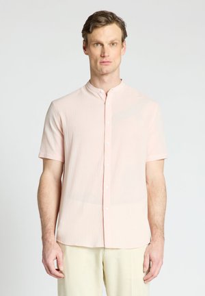 Homme portant une chemise rose clair à manches courtes avec col mao et un pantalon couleur crème, debout devant un fond blanc uni.