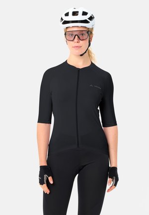 Femme portant un maillot de cyclisme noir, des gants rembourrés noirs, un casque blanc et des lunettes de protection, debout devant un fond clair uni.