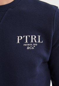 Felpa blu navy con tessuto testurizzato, colletto a girocollo e logo stampato bianco che recita "PTRL PETROL IND & Co." sul petto.