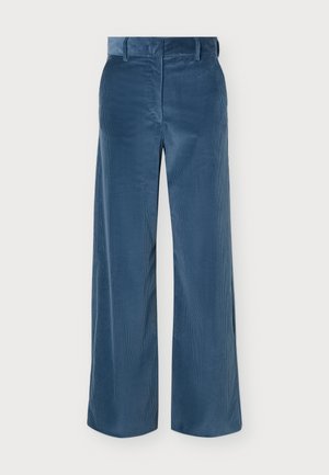 Mujer que lleva una camisa abotonada estampada con mangas a rayas metida en pantalones anchos de talle alto azules y zapatos negros, de pie mirando hacia adelante.