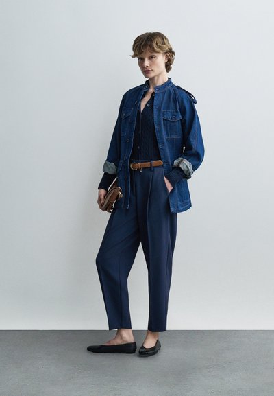 Person iført en mørkeblå denimjakke over en sort sweater, marineblå bukser med et brunt bælte, sorte flade sko og holder en lille brun clutch.