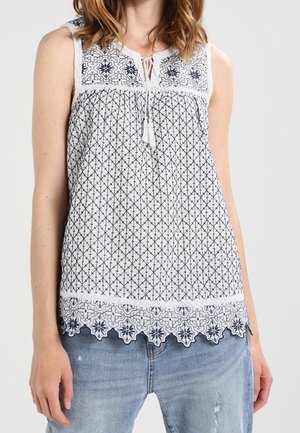 Mouwloze witte blouse met zwarte geometrische en bloemrijke borduurwerk, kwastje aan de halslijn, gedragen met lichtblauwe versleten spijkerbroek.