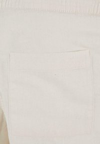 Détail de la poche arrière sur un pantalon beige clair en lin ou coton, montrant la texture du tissu et les coutures.