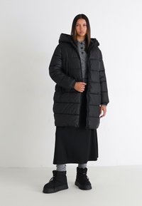 Svart boblejakke med hette, quiltet design og frontlukking. Båret over grå topp og svart skjørt med kraftige svarte støvler.
