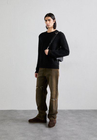 Pull en tricot noir texturé, jeans bruns déchirés et chaussures en suede marron. Modèle portant un sac bandoulière avec une sangle noire. Fond neutre.