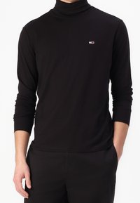 Black turtleneck long-sleeve shirt in soft fabric, featuring a small embroidered logo in red and white on the chest. Tailored fit.