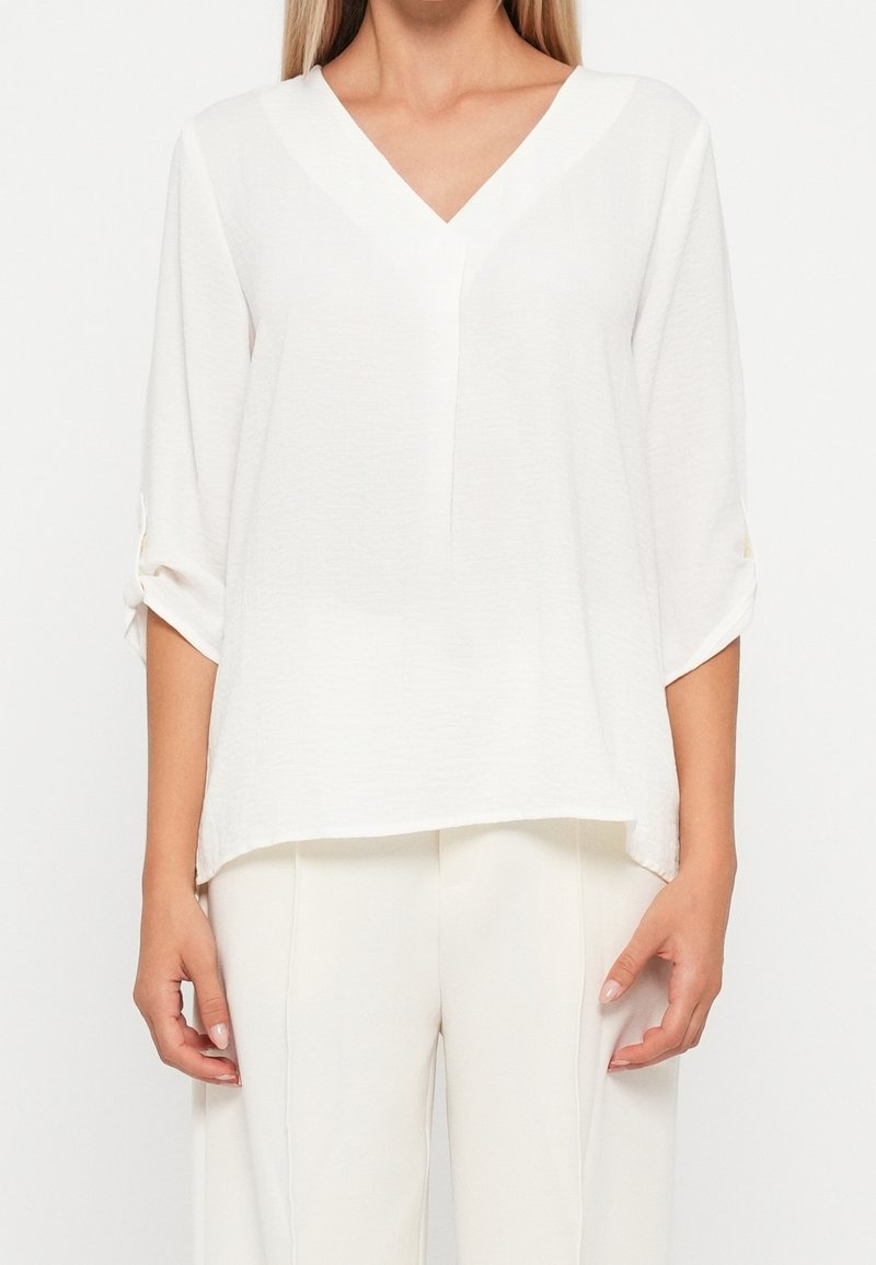 Femme portant un chemisier blanc à col en V avec manches 3/4 retroussées et pantalon blanc, debout sur un fond blanc uni.