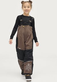 Bruine en zwarte sneeuwbroeken met verstelbare bandjes, grote zakken en een geometrisch logo op het been, gedragen over een zwart longsleeve shirt.