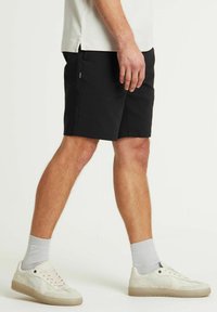 Zwarte shorts van lichtgewicht materiaal, met zijzakken en een rechte snit. Draagt lichtgekleurde sneakers en grijze sokken.