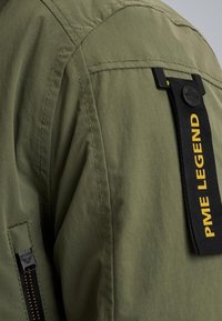 Olijfgroen jack van een gestructureerde stof, met een zwart label met de tekst "PME LEGEND" en een zijvak met rits en gouden hardware.