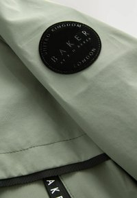 Lichtgroene jassenstof met een matte afwerking, voorzien van een zwart rond patch met het label "BAKER" in witte letters en logo aan de voorkant.