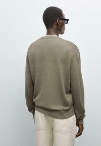 Übergröße olivgrüner Pullover aus weichem Strickstoff mit gerippten Bündchen und Saum. Über hellen Hosen getragen, von hinten betrachtet.