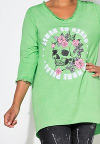 Chemise verte à manches longues avec un col et des manches effilochés, ornée d'un graphique de crânes et de roses, accompagnée de texte. Coupe décontractée et confortable.