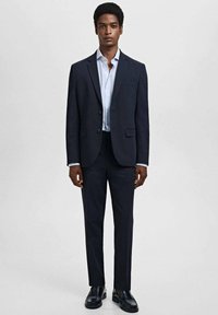 Donker marineblauw pak met een enkelvoudig gesloten jasje en bijpassende broek. Lichte stof met een gladde textuur; gecombineerd met een lichtblauw overhemd.