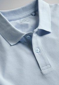 Polo en coton bleu clair avec col et deux boutons, affichant l'étiquette "THE SET Fabriqué au Bangladesh."