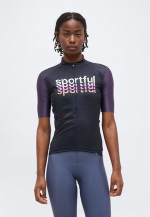 Γυναίκα με μαύρο και μοβ ποδηλατικό μπλουζάκι με την επιγραφή «sportful» και ταιριαστόι επενδεδυμένοι ποδηλατικοί σορτς, στέκεται σε λευκό φόντο.