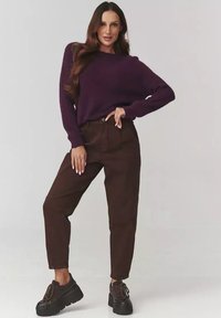 Kobieta pozująca w ciemnofioletowym swetrze, ciemnobrązowych spodniach o wysokim stanie i czarnych platformowych butach na sznurówki na jednolitym tle.