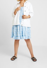 Weiße Jeansjacke über einem blauen Batikkleid mit gerafftem Saum und schwarzen Sandalen mit silberner Hardware. Leichter, lässiger Stoff.