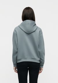 Personne aux cheveux lisses et foncés portant un sweat à capuche gris clair et un pantalon noir, vue de dos sur un fond blanc uni.