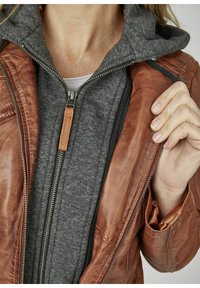 Giacca di pelle marrone sopra una felpa grigia con zip. La giacca ha una finitura testurizzata e zip a vista con un tirante in pelle beige.